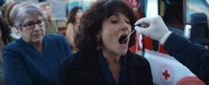 A woman opens her mouth wide for a DNA swab. A line of people behind her await their turn.